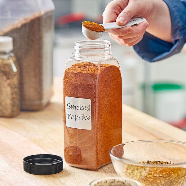 A person pouring spice into a rectangular plastic container with a black lid.