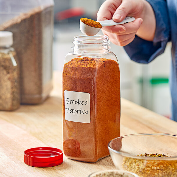 A person pouring smoked paprika into a rectangular plastic spice container with a flat red lid.