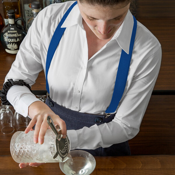 A woman in white shirt and blue Henry Segal suspenders pouring a drink into a glass.
