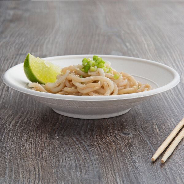 A white compostable sugarcane noodle bowl with noodles, green onions, and a lime wedge on a wood surface with chopsticks.