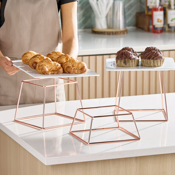 A three-piece set of square display stands with rose gold metal frames and flat surfaces for displaying food or items.