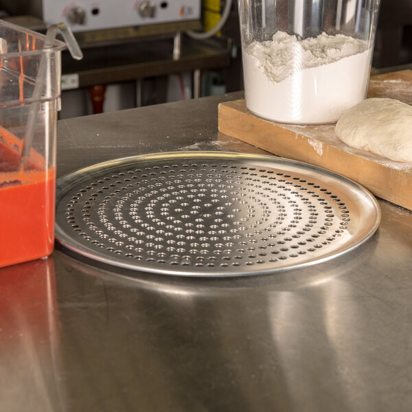 An American Metalcraft aluminum pizza pan with perforations holding pizza dough on a counter.