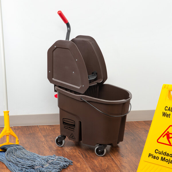 A Rubbermaid brown mop bucket with a down press wringer next to a mop on the floor.