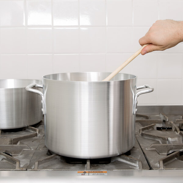 A person stirring a Vollrath Arkadia aluminum stock pot on a stove with a wooden spoon.