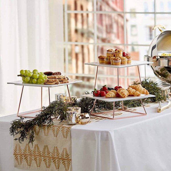 A table with Acopa rose gold wire display risers holding trays of food and fruit.