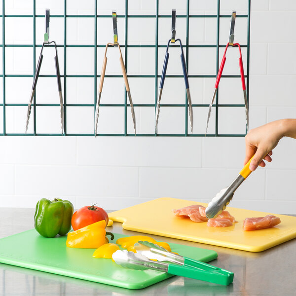 A person using Vollrath tongs with green, yellow, and red accents to cut vegetables on a cutting board.