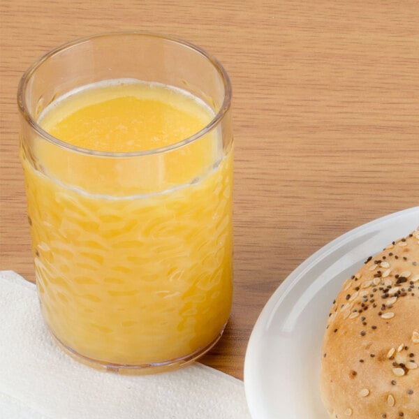 A Carlisle clear plastic tumbler filled with orange juice next to a plate of bread with a bagel on it.