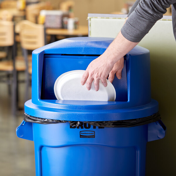 A person putting a plate into a blue Rubbermaid BRUTE trash can lid.