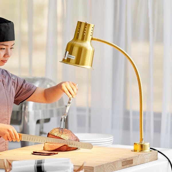 A woman cutting a piece of meat on a wooden cutting board with an Avantco gold heat lamp overhead.