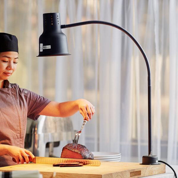 A woman using a fork and tongs to cut meat under an Avantco black countertop heat lamp.