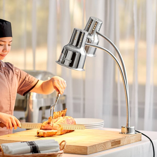 A woman cutting meat on a wooden cutting board with an Avantco stainless steel heat lamp overhead.