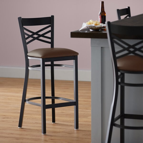 A Lancaster Table & Seating black metal cross back bar stool with a dark brown vinyl seat.