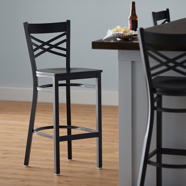 A Lancaster Table & Seating black cross back bar stool with a black wood seat next to a table.