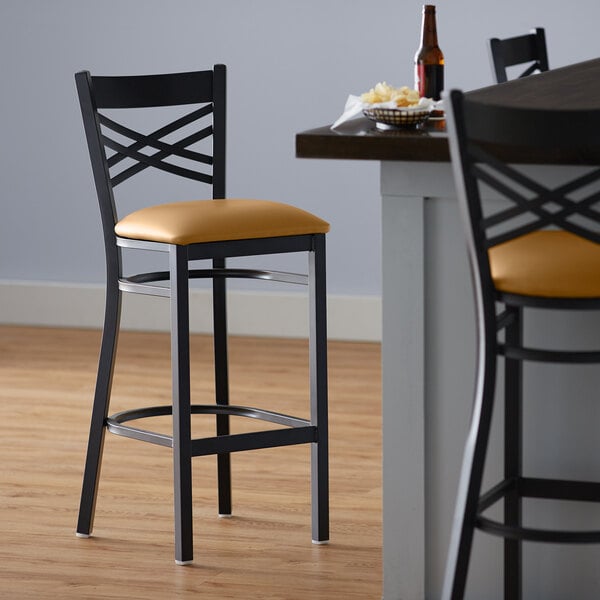 A Lancaster Table & Seating black cross back bar stool with light brown vinyl padding.