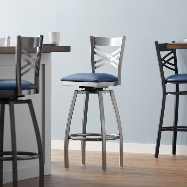 A Lancaster Table & Seating silver cross back swivel bar stool with a navy blue cushion.