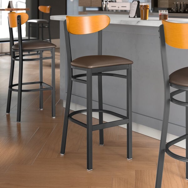 A group of Lancaster Table & Seating Boomerang Series bar stools with dark brown vinyl seats and cherry wood backs next to a counter.