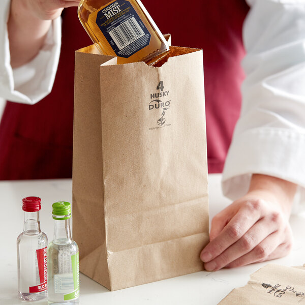 A person's hand putting a clear bottle of alcohol into a Duro brown paper bag with black text.