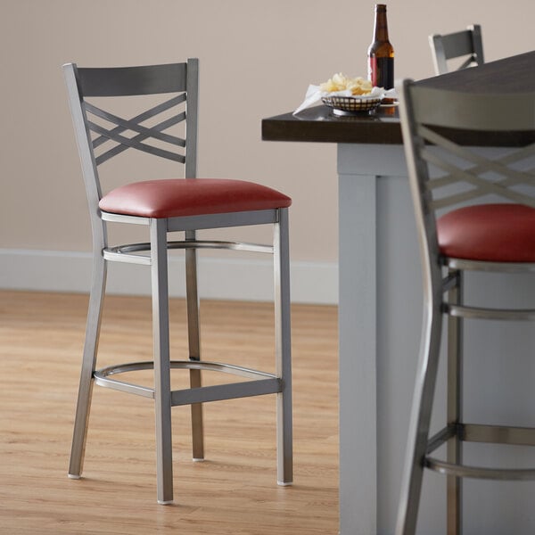 A Lancaster Table & Seating cross back bar stool with a red vinyl padded seat next to a table.