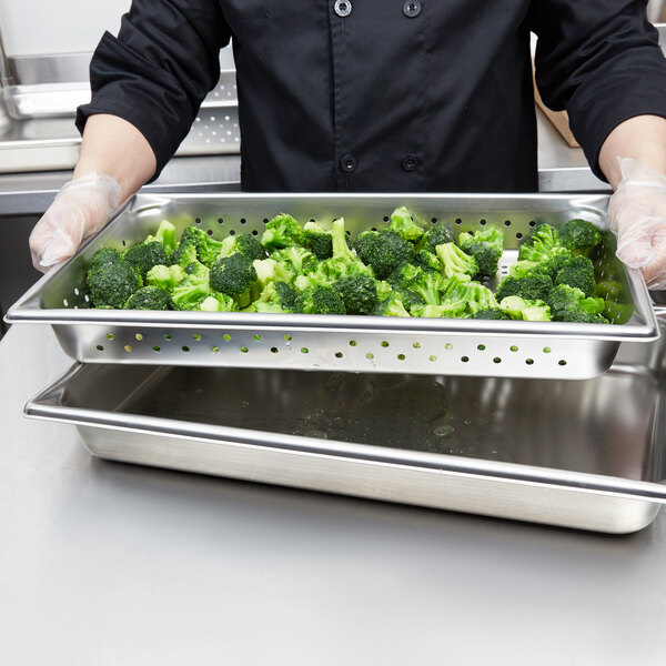 A person in a chef's uniform holding a Vollrath stainless steel tray of broccoli.