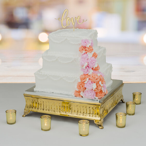 A Tabletop Classics gold-plated square cake stand with a white cake topped with flowers displayed on a table.