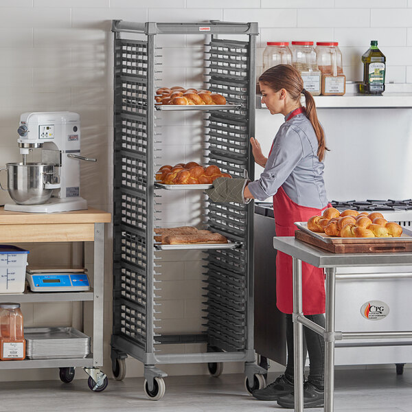 A woman in a red apron standing in front of a Cambro sheet pan rack full of doughnuts in a bakery.