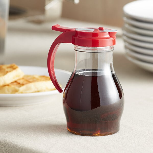 A clear polycarbonate teardrop pitcher of syrup with a red top.