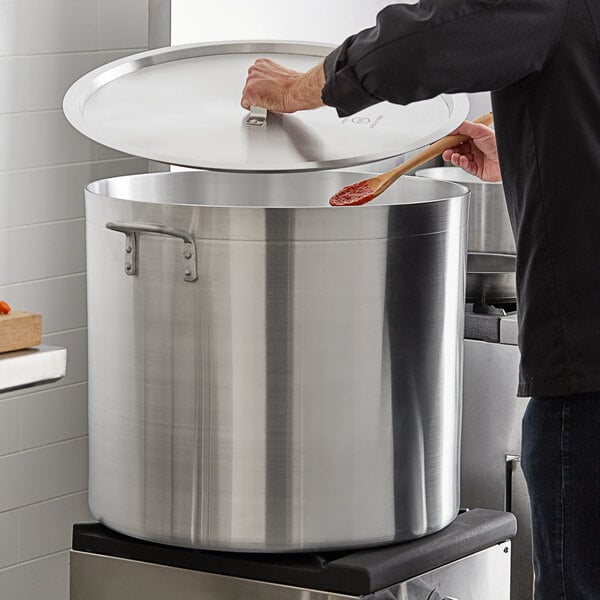 A person holding a wooden spoon over a large silver pot with a black and silver lid.