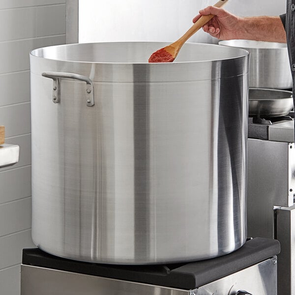 A man stirring a large Choice aluminum stock pot on a stove in a professional kitchen.