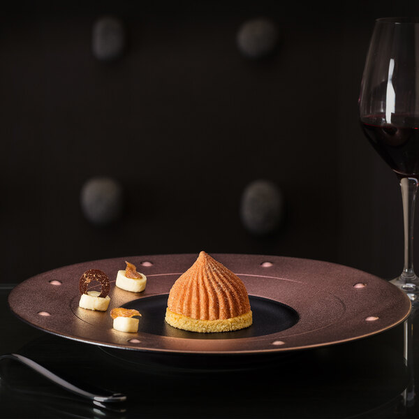 A black and bronze RAK Porcelain round plate with a dessert on it on a table with a glass of wine.