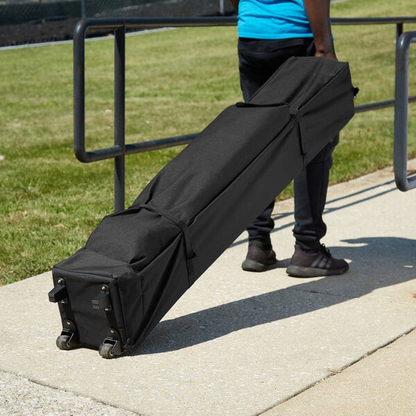 A person carrying a black Backyard Pro canopy roller bag on a sidewalk.