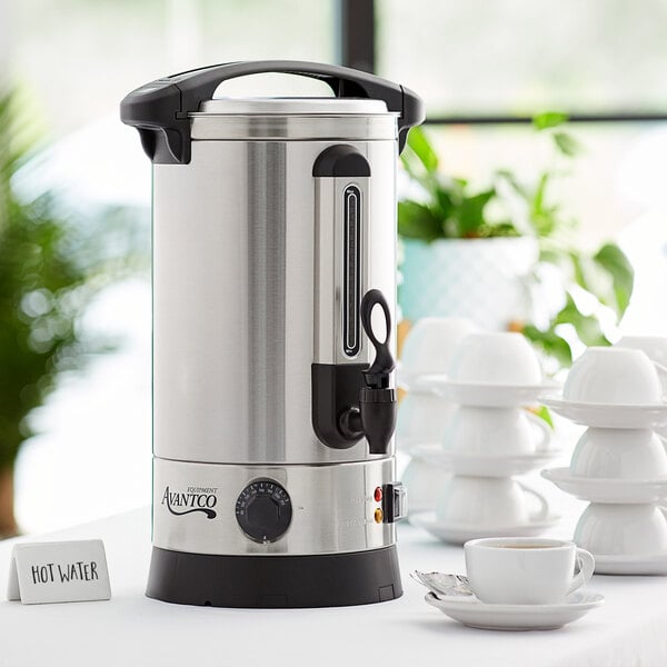 An Avantco silver and black water boiler on a table with a cup and saucer.