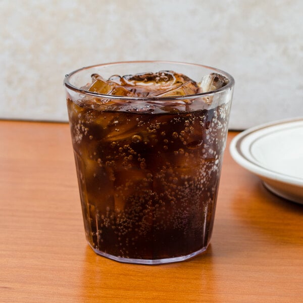 A Cambro clear plastic tumbler filled with soda and ice on a table with a plate of food.