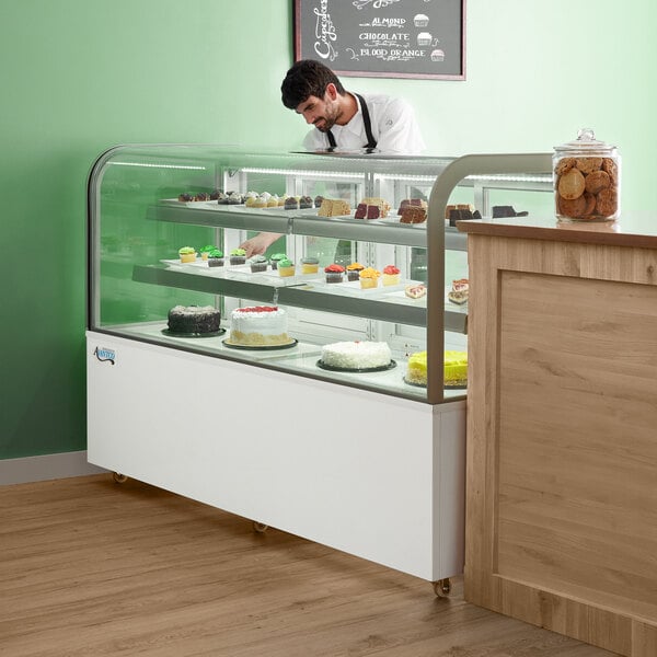 A man in a white shirt and black suspenders looking at an Avantco refrigerated bakery display case filled with cakes.