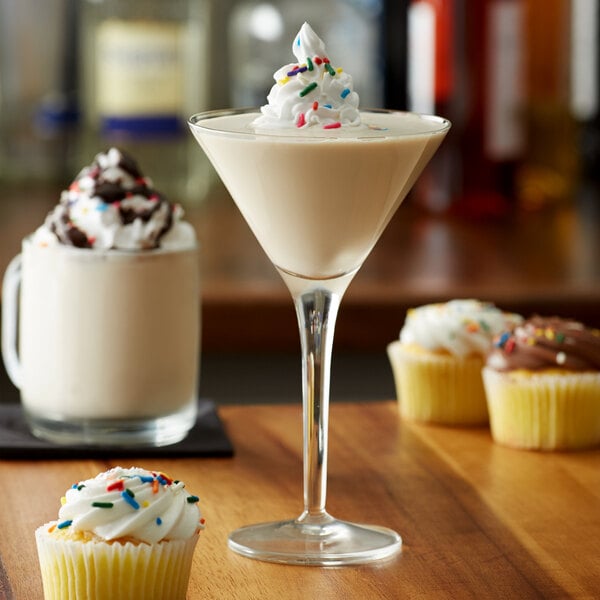A close-up of a cupcake with white frosting and sprinkles on a table with a glass of Monin Premium Cupcake Flavoring syrup.