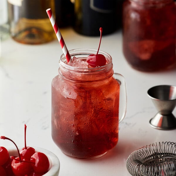 A glass jar with Monin Premium Tart Cherry Flavoring Syrup in a red drink with ice and a straw.