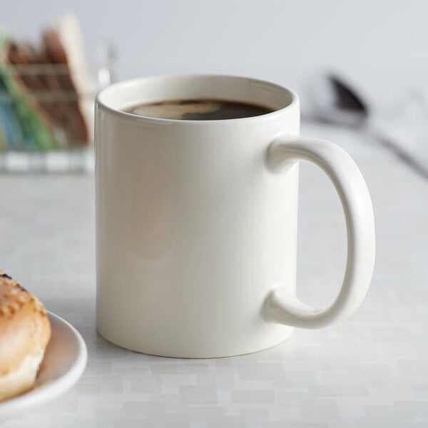 An Acopa ivory stoneware mug filled with brown liquid next to a donut on a plate.