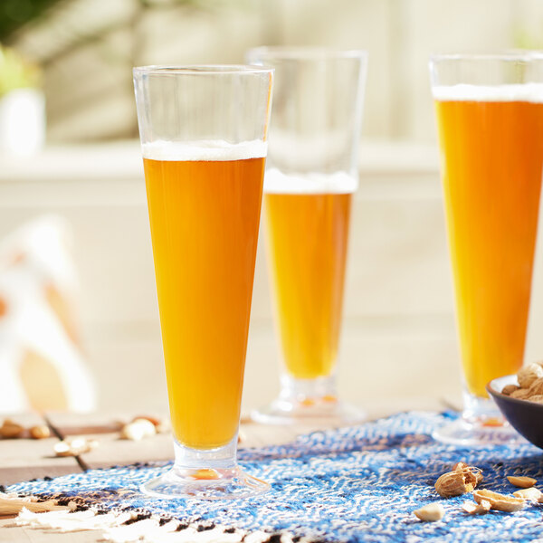 Three Libbey Tritan plastic footed pilsner glasses filled with beer on a table with nuts.