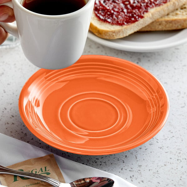 A hand holding a cup of coffee over a Tuxton papaya saucer on a table with a plate and bread.
