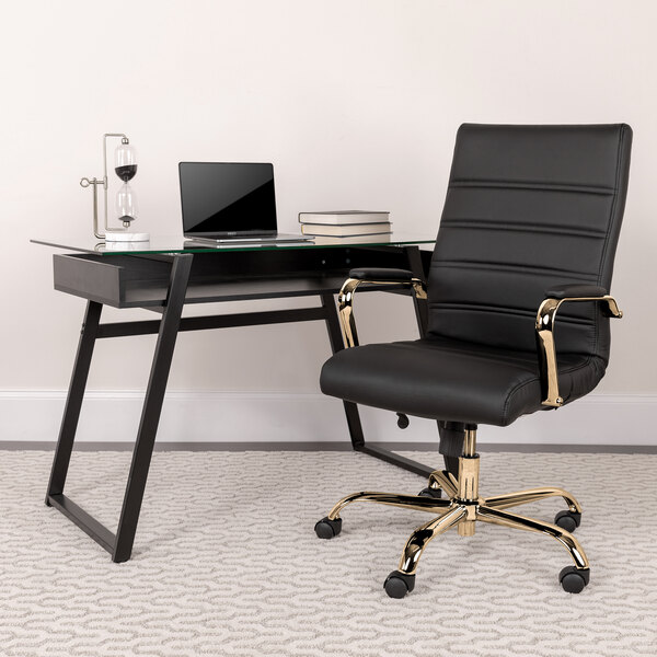 A Flash Furniture black leather office chair with gold arms and base next to a glass desk with a laptop on it.