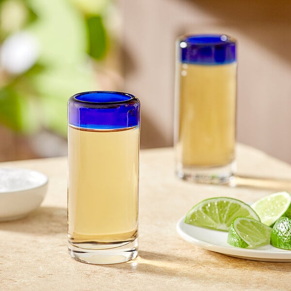 Two Acopa shot glasses with blue rims filled with tequila next to a lime wedge.