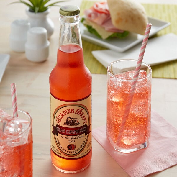 A bottle of Reading Soda Works Cran Raspberry soda next to a glass of pink liquid with a straw.