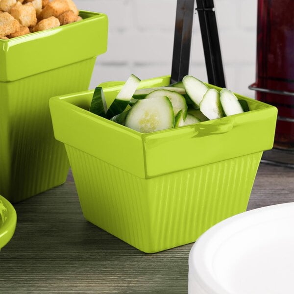 A lime green square Tablecraft condiment bowl with cucumbers and crackers inside.