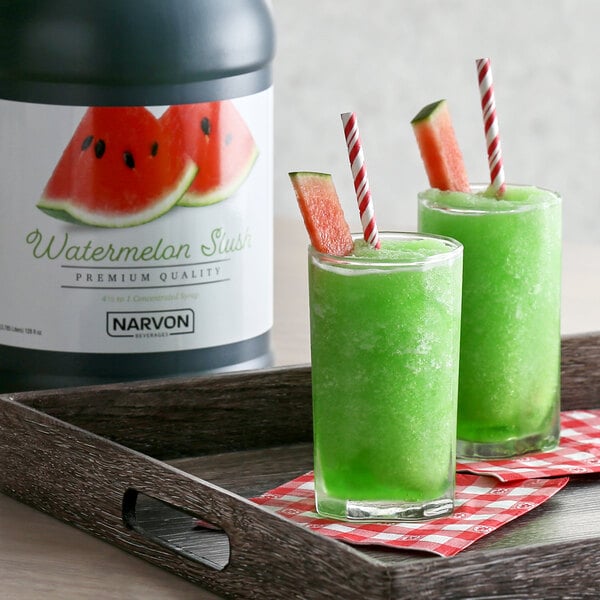 A close-up of a container of Narvon Watermelon Slushy Concentrate with a glass of green liquid and a straw on a tray.