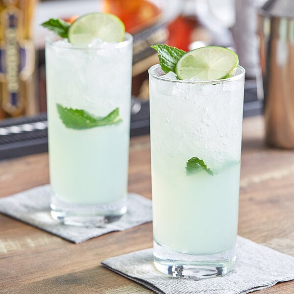 Two Acopa beverage glasses of liquid with ice and lime slices on a table with lime and mint garnish.