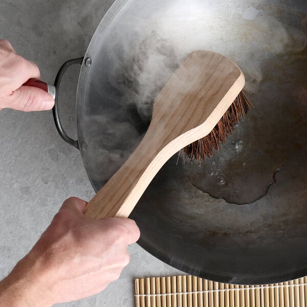 A person using a Thunder Group Palmyra bristled wok brush with a wood handle to clean a wok.
