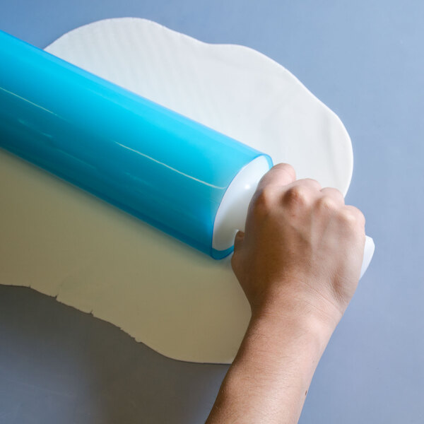 A hand rolling a blue roll of plastic over a white table using a blue Ateco silicone rolling pin cover.