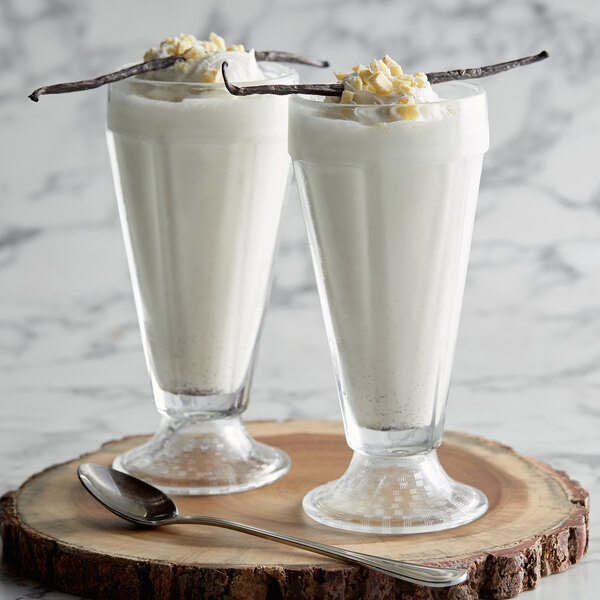 Two glasses of vanilla milkshakes with vanilla sticks on a wood stand.
