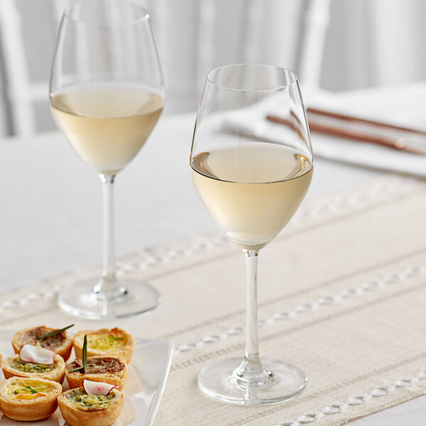 Two Acopa wine glasses filled with white wine next to a plate of food.