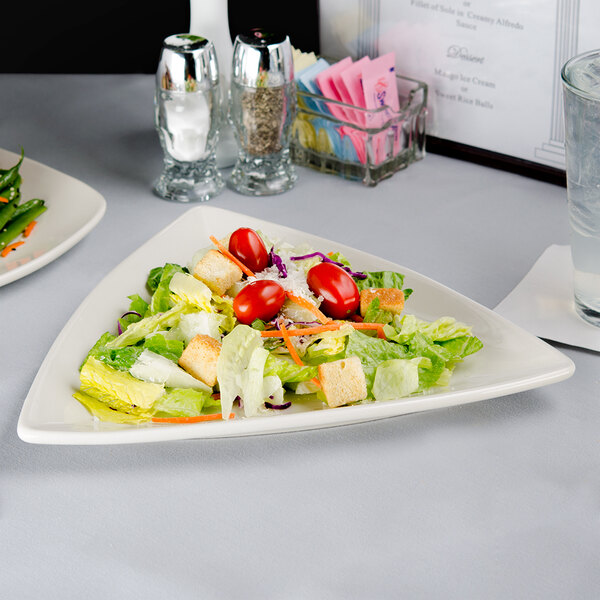 A Tuxton eggshell triangle china plate with a salad on a table.