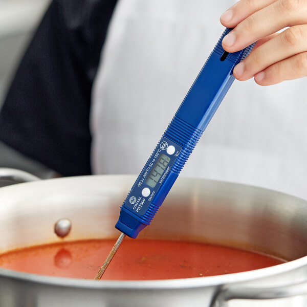 A hand holding a Comark digital pocket probe thermometer in a pot of soup.